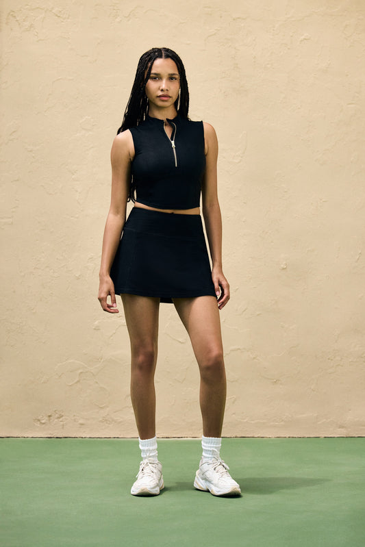 Tennis Club black ribbed knit skirt with built-in shorts worn by a model with a matching zip-up top on a tennis court